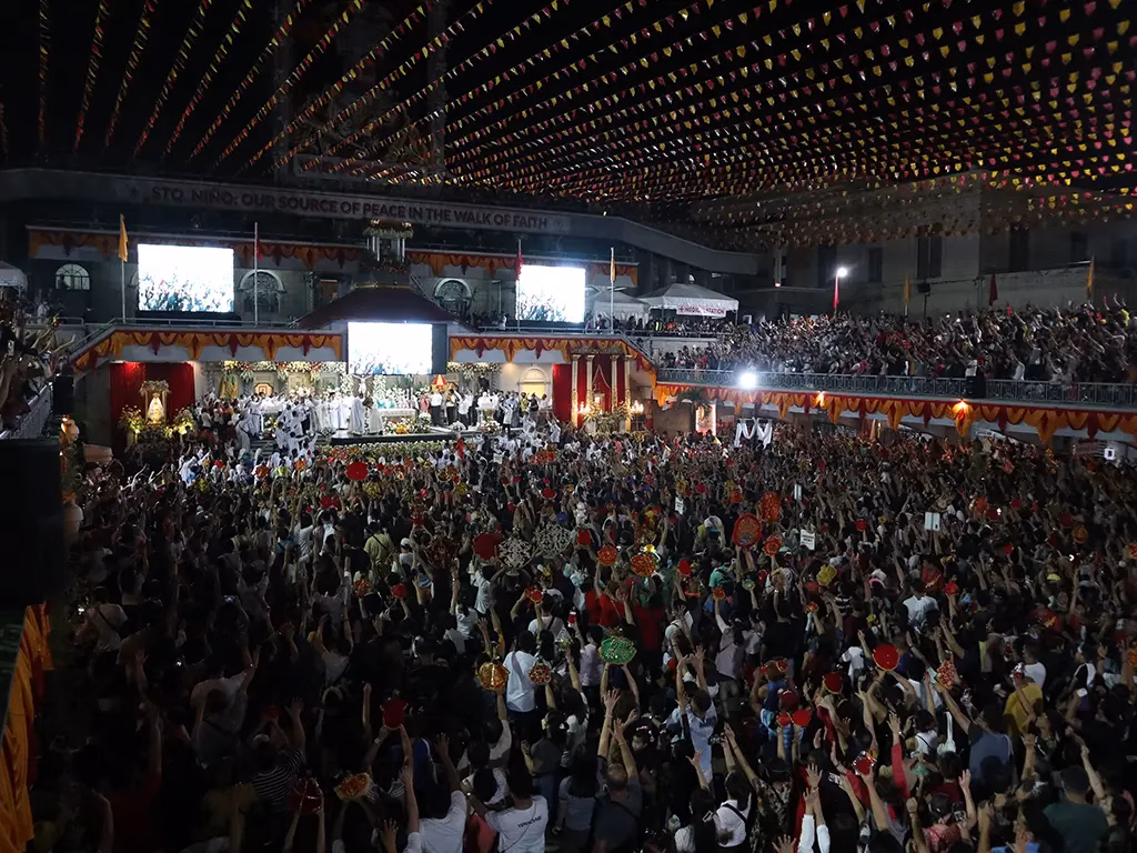 Thousands of devotees with arms raised fill the grounds of the Basilica Minore del Santo Niño de Cebu during the Sinulog Festival novena in Cebu City, Philippines, holding decorated Santo Niño statues aloft beneath colorful festival bunting during a nighttime outdoor mass.