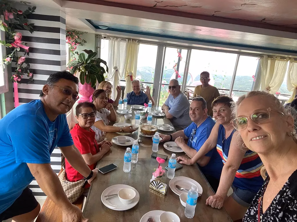 A group of family and friends gathered around a long dining table inside Cafe Alicia, with the café's distinctive striped walls, pink floral décor, and large windows overlooking a green hillside landscape visible in the background.