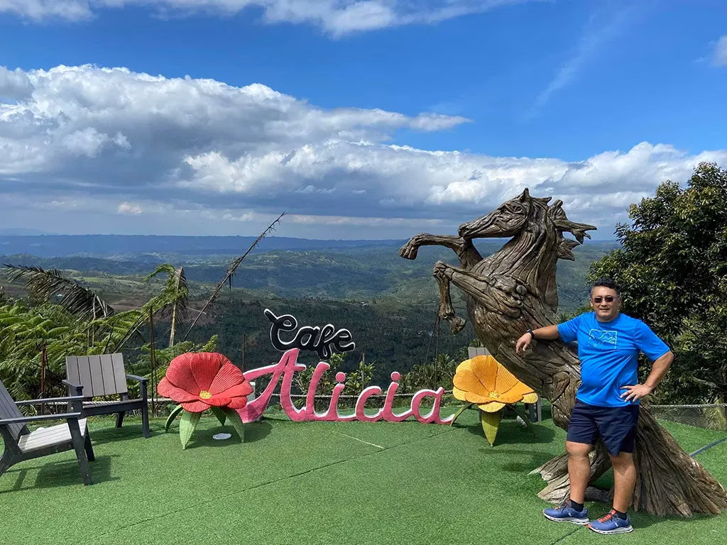The outdoor viewing deck of Cafe Alicia featuring a large driftwood horse sculpture and pink floral signage, with a visitor posing beside it and a sweeping panoramic view of the green highlands of Negros Oriental stretching to the horizon under a dramatic sky.