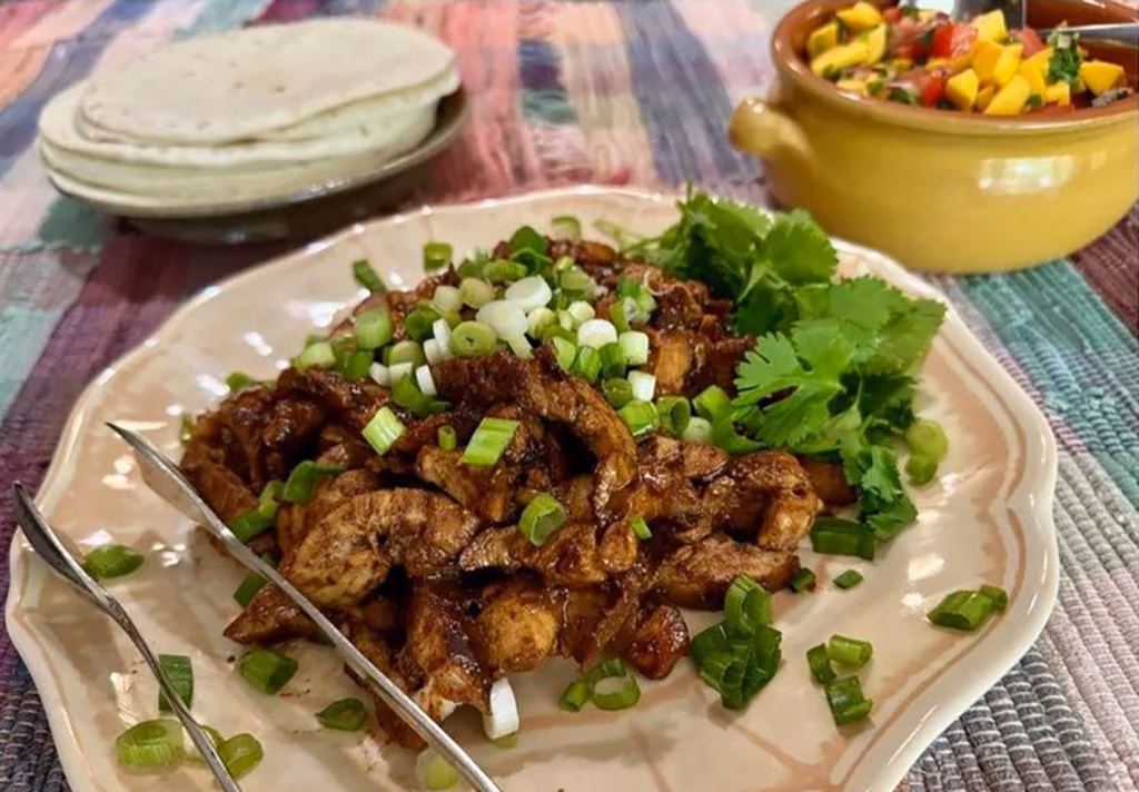 A serving plate of Filipino chicken adobo prepared as taco filling, topped with sliced green onions and fresh cilantro, served alongside a stack of flour tortillas and a yellow bowl of mango salsa on a colorful placemat.