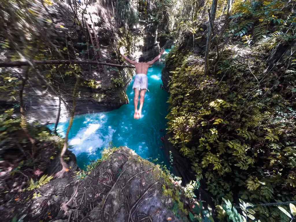 A picture of someone diving from a cliff into the water about 50 feet below at Kawasan Falls, in the Philippines.