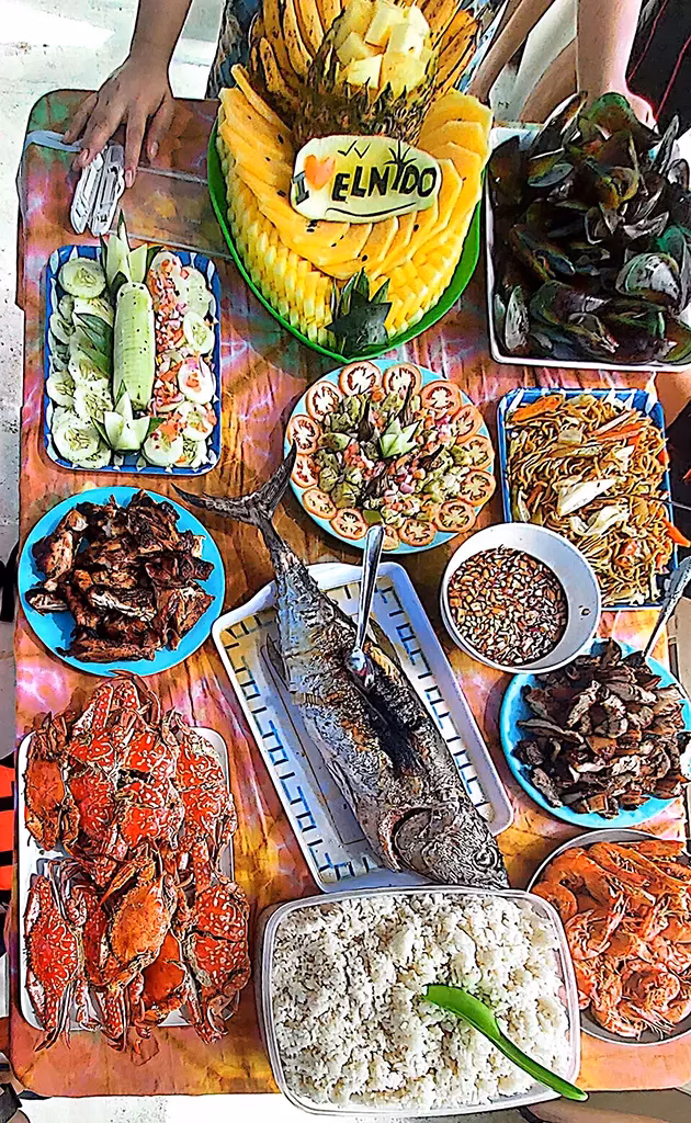 An overhead view of a traditional Filipino boodle feast spread in El Nido, Palawan, Philippines, featuring whole grilled fish, spicy crabs, shrimp, mussels, grilled meat, pancit noodles, rice, fresh salad, and a carved pineapple centerpiece reading "I Love El Nido."