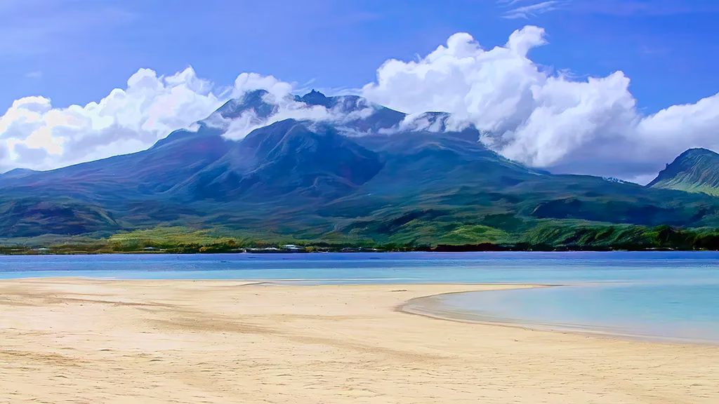 A white sandbar curving into turquoise water in the foreground with Mount Hibok-Hibok volcano rising dramatically behind it on Camiguin Island, Philippines, its upper slopes partially shrouded in clouds against a vivid blue sky.