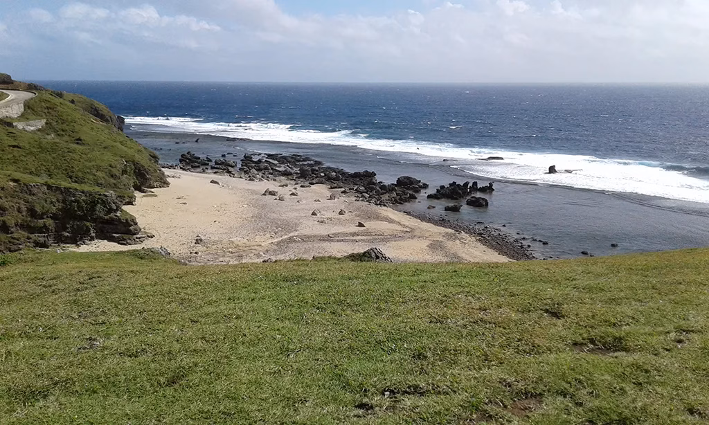 A rugged coastline in Batanes, Philippines, viewed from a grassy clifftop, showing a rocky cove with dark sand, powerful surf breaking along the shoreline, deep blue open ocean stretching to the horizon, and a coastal road visible on the headland to the left.