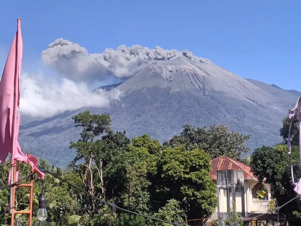Kanlaon Volcano erupting with a large ash column rising from its summit against a clear blue sky, viewed from a nearby town in Negros, with residential buildings, tropical trees, and pink festival flags visible in the foreground.