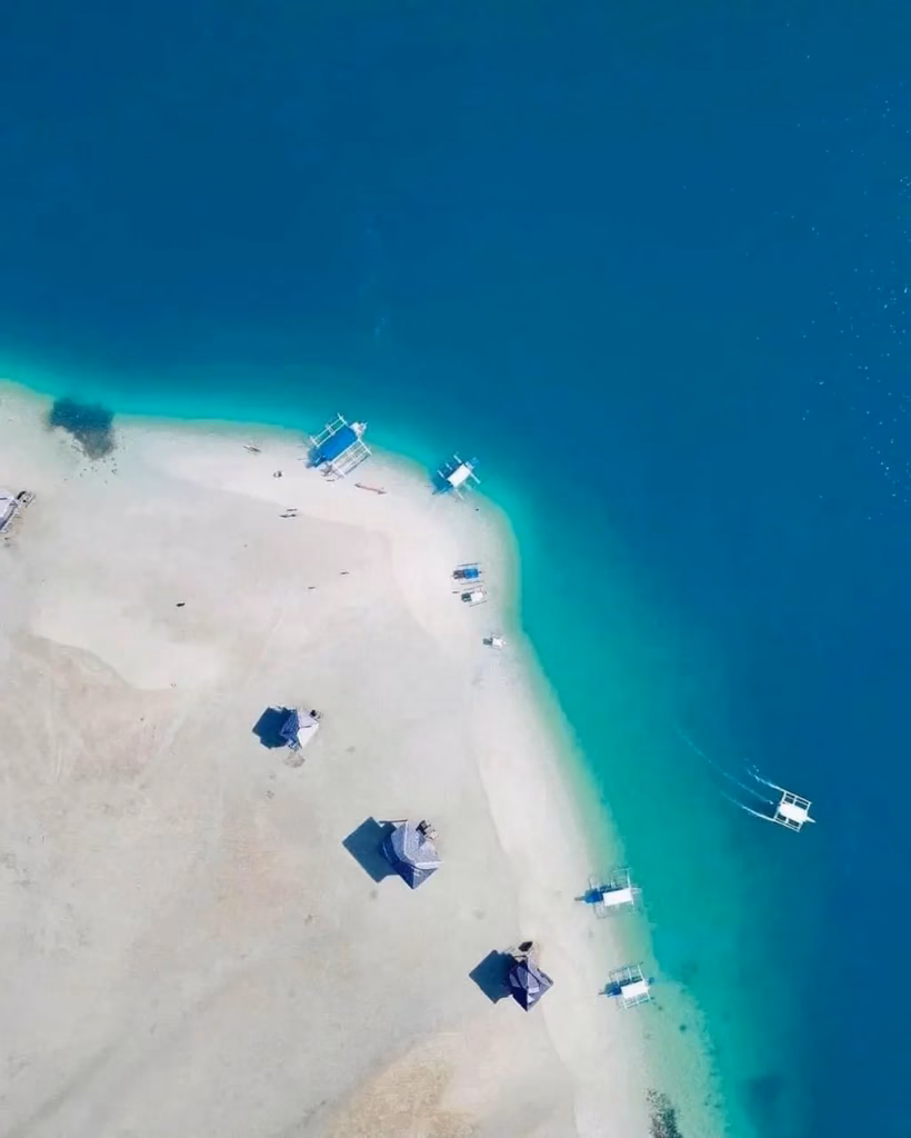 Aerial view of Manjuyod White Sandbar in Negros Oriental, Philippines, showing a broad white sand bank curving through vivid turquoise and deep blue water, with small nipa cottage structures and bangka boats scattered along its edges.
