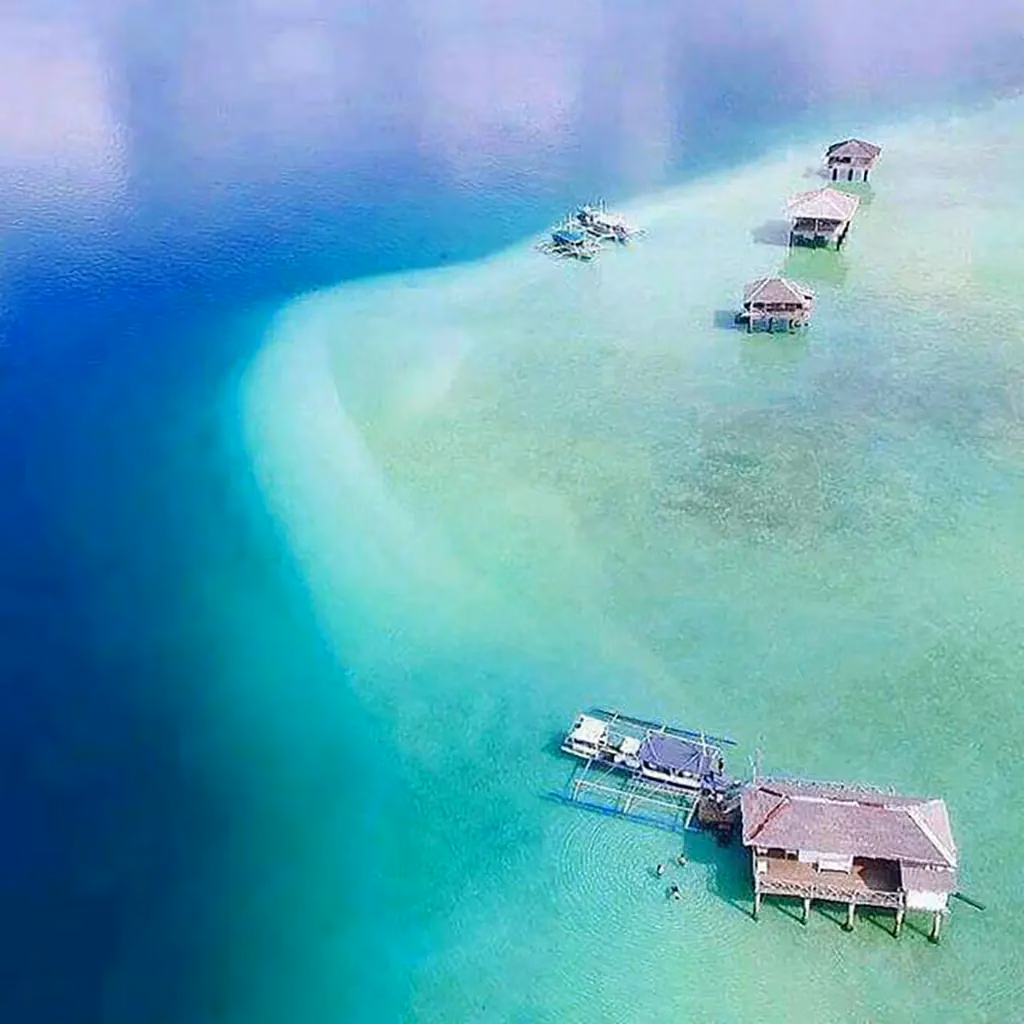 Aerial view of overwater nipa cottages at Manjuyod White Sandbar in Negros Oriental, Philippines, partially submerged at high tide, surrounded by a vivid gradient of deep blue, turquoise, and jade green water revealing the sandbar's shape beneath the surface.