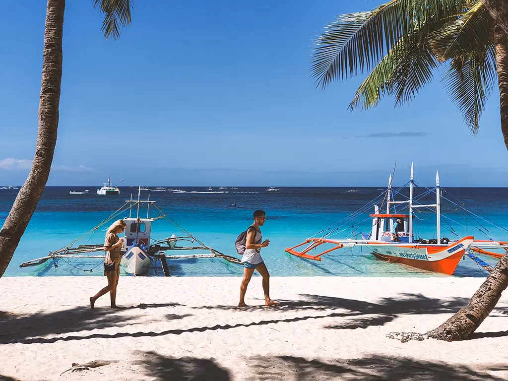 Tourists walkin along White Beach in Boracay with a couple of banca's moored on the shore.