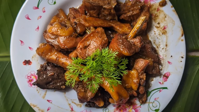 A plate of Filipino Adobo sa Gata, chicken pieces braised in vinegar, garlic, and coconut milk until deeply glazed, garnished with fresh green herbs and served on a floral-rimmed white plate atop a banana leaf.