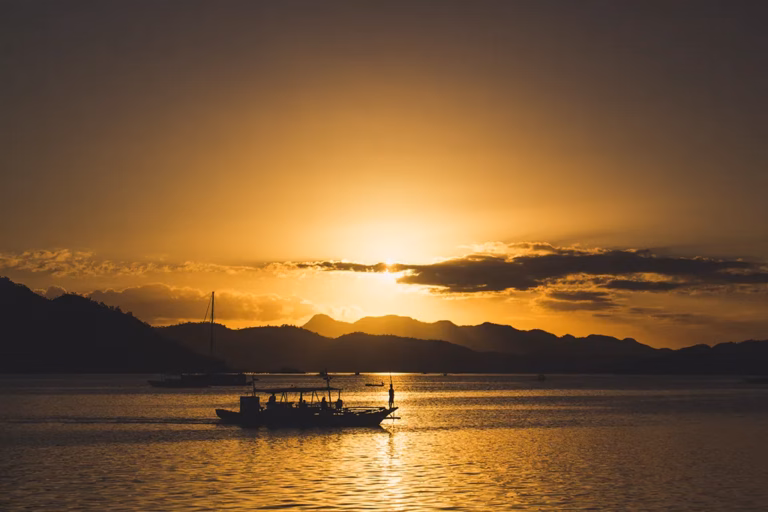 A silhouetted bangka boat with a figure standing at the bow crosses the golden waters of Coron Bay at sunset in Palawan, Philippines, with the mountain ridgeline of Busuanga Island and dramatic clouds glowing amber and orange against the evening sky.