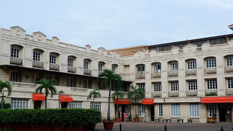 The Spanish colonial facade of Casa Manila museum in Intramuros, Manila, featuring cream-colored walls, red canvas awnings, wrought-iron balconies, and palm trees in the courtyard.