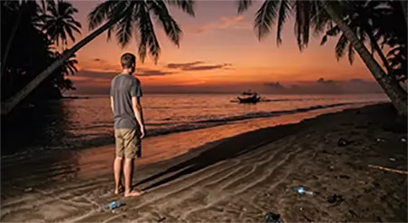 Stranded traveler watching a Bangka motor away from a remote Philippine island beach at sunset, island hopping in the Philippines gone wrong